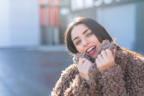 Lovely young woman smiling and looking at camera while wrapping in warm stylish coat and standing on blurred background of city street on sunny day — Stock Photo