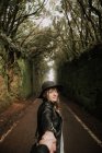 Side view of elegant lady in hat and leather jacket holding hand of person and standing on footpath between murk alley of high walls and woods — Stock Photo