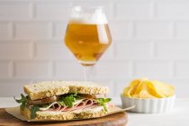 Delicious sandwich with ham, cheese and greens with glass of beer and chips on white background — Stock Photo