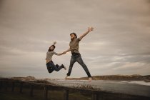 Junge glückliche Frau und Mann mit Hüten halten Händchen und haben Spaß auf Sitz an der Küste in der Nähe von winkendem Meer und wolkenverhangenem Himmel — Stockfoto