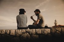 Вид збоку стильний хлопець в капелюсі курильної труби і елегантна жінка в капелюсі читає гучність і сидить на каменях біля маяка — стокове фото