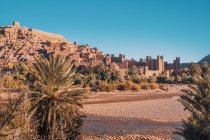 Construcciones rocosas en la ciudad vieja cerca de árboles verdes en la orilla del río y el cielo azul en Marrakech, Marruecos - foto de stock