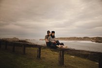 Junger Mann mit Hut neben eleganter Frau, die auf Handtrommel in Mütze auf Sitz in der Nähe der Küste von Meer und bewölktem Himmel spielt — Stockfoto