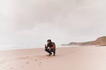 Man in warm wear with backpack smoking pipe on sand coast near sea and sky in clouds — Stock Photo