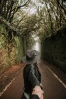 Side view of elegant lady in hat and leather jacket holding hand of person and standing on footpath between murk alley of high walls and woods — Stock Photo
