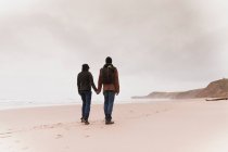 Back view of young couple with backpack in warm wear holding each other hands on sand beach near sea and hills — Stock Photo