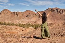 Vista posterior de la joven morena de pie entre tierras desiertas cerca de antiguas construcciones y colinas en Marrakech, Marruecos - foto de stock