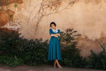 Donna in abito blu con le mani incrociate in piedi vicino a piante verdi che crescono vicino muro di pietra invecchiato — Foto stock
