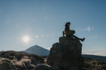 Seitenansicht einer unkenntlichen Reisenden, die an sonnigen Tagen auf einem Felsen am Hang sitzt — Stockfoto