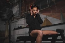 Seitenansicht einer Frau in Sportbekleidung mit Kopfhörern und Gadget, die Musik hört und im Fitnessstudio sitzt — Stockfoto