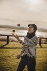 Elegant woman in cap juggling balls on grass near coast of sea and sky with sun — Stock Photo