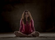 Jovem cansada senhora em vestido vermelho sentado em pose de lótus entre a areia e olhando para a câmera no quarto escuro — Fotografia de Stock