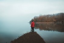 Rückansicht des Fotografen im roten Mantel, der am Ufer des ruhigen Sees im Nebel steht — Stockfoto