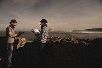 Élégant gars en chapeau et femme élégante en bouchon de lecture des volumes près des rochers sur la côte de la mer et le ciel merveilleux — Photo de stock