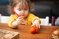 Menina adorável criança usando pincel para pintar ovo de Páscoa enquanto sentado à mesa — Fotografia de Stock