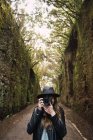 Elegant lady in hat and leather jacket shooting on camera and standing on footpath between murk alley of high walls and woods — Stock Photo
