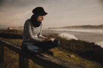 Elegant woman in cap taking notes in notepad and sitting on seat on coast near waving sea — Stock Photo