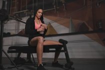 Femme en vêtements de sport vérifier l'heure sur la montre et assis sur le banc dans la salle de gym — Photo de stock