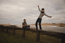 Young happy woman and man in hats having fun on seat on coast near waving sea and cloudy sky — Stock Photo
