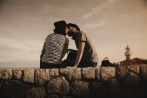 Vista laterale di elegante elegante coppia baciare e seduto su rocce vicino faro e cielo meraviglioso — Foto stock