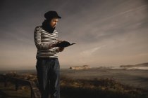 Елегантна жінка в кепці приймає нотатки в блокноті і сидить на сидінні на узбережжі біля махаючого моря — стокове фото