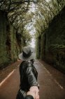 Vista lateral de señora elegante en sombrero y chaqueta de cuero cogida de la mano de la persona y de pie en el sendero entre callejón oscuro de paredes altas y maderas - foto de stock