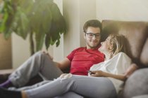 Homme et femme joyeux couché et assis sur le canapé tout en tenant la télécommande et regarder la télévision — Photo de stock