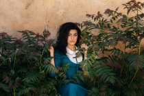 Beautiful young brunette Moroccan woman in blue gown standing between green plants growing near aged stone wall — Stock Photo