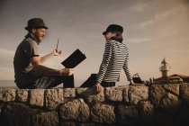 Side view of stylish guy in hat smoking pipe and elegant woman in cap reading volume and sitting on rocks near beacon — Stock Photo