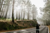 Donna che scatta foto in una strada asfaltata nella foresta nebbiosa con tronchi d'albero alti ricoperti di muschio — Foto stock