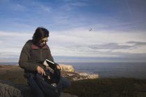 Mujer elegante sacando un libro de la mochila y sentado en el prado cerca de la baliza en la orilla y el cielo maravilloso - foto de stock