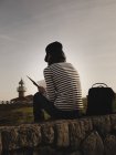 Rückansicht einer eleganten Frau mit Mütze und Leseband, die auf einer Wiese in der Nähe von Leuchtturm am Ufer und herrlichem Himmel sitzt — Stockfoto