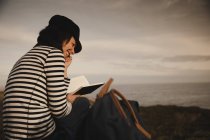 Visão traseira da mulher elegante no volume de leitura do boné e sentado no prado perto do farol na costa e no céu maravilhoso — Fotografia de Stock