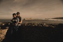 Cara elegante em chapéu e mulher elegante em volume de leitura de boné perto de rochas na costa do mar e céu maravilhoso — Fotografia de Stock