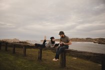 Junger Mann mit Hut spielt auf Handtrommel neben eleganter Frau in Mütze mit Notizblock und Bleistift auf Sitz in der Nähe der Küste — Stockfoto