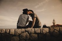 Seitenansicht von stilvollen eleganten Paar küsst und sitzt auf Felsen in der Nähe von Leuchtfeuer und wunderbaren Himmel — Stockfoto