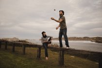Young guy in hat juggling balls near elegant woman in cap with ethic drum sitting on seat near coast of sea and cloudy sky — Stock Photo