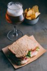 Delicioso sanduíche com presunto, queijo e verduras com copo de cerveja e batatas fritas no fundo escuro — Fotografia de Stock
