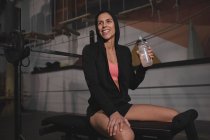 Femme en vêtements de sport avec bouteille d'eau assise sur le banc dans la salle de gym — Photo de stock