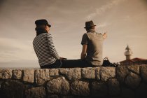 Вид збоку на стильну елегантну пару, що сидить на каменях біля маяка і чудове небо — стокове фото