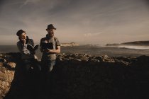 Стильный парень в шляпе и элегантная женщина в кепке читают тома у скал на берегу моря и чудесного неба — стоковое фото