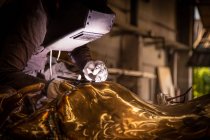 Людина в зварювальному шоломі, що працює з металевим виробом на маленькому ливарному заводі — стокове фото