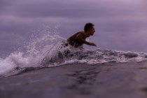 Vue latérale du mâle flottant sur la planche de surf entre l'eau de mer et le ciel nuageux sur Bali, Indonésie — Photo de stock