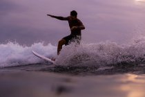Hombre surfeando entre olas de agua de mar con salpicaduras y cielo nublado por la noche en Bali, Indonesia - foto de stock