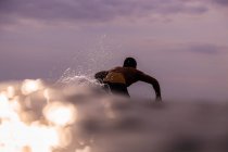 Vue arrière du mâle flottant sur la planche de surf entre l'eau de mer et le ciel nuageux sur Bali, Indonésie — Photo de stock