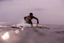 Visão traseira do macho flutuando na prancha de surf entre a água do mar e céu nublado em Bali, Indonésia — Fotografia de Stock
