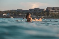 Side view of cheerful female floating on surf board between water of sea and blue heaven on Bali, Indonesia — Stock Photo