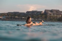 Vista lateral da fêmea alegre flutuando na prancha de surf entre a água do mar e o céu azul em Bali, Indonésia — Fotografia de Stock