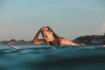 Vista laterale di allegra femmina galleggiante su tavola da surf tra acqua di mare e cielo blu a Bali, Indonesia — Foto stock