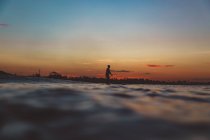 Vista lateral da silhueta de macho com pá na prancha de surf entre a água do mar e do céu à noite em Bali, Indonésia — Fotografia de Stock